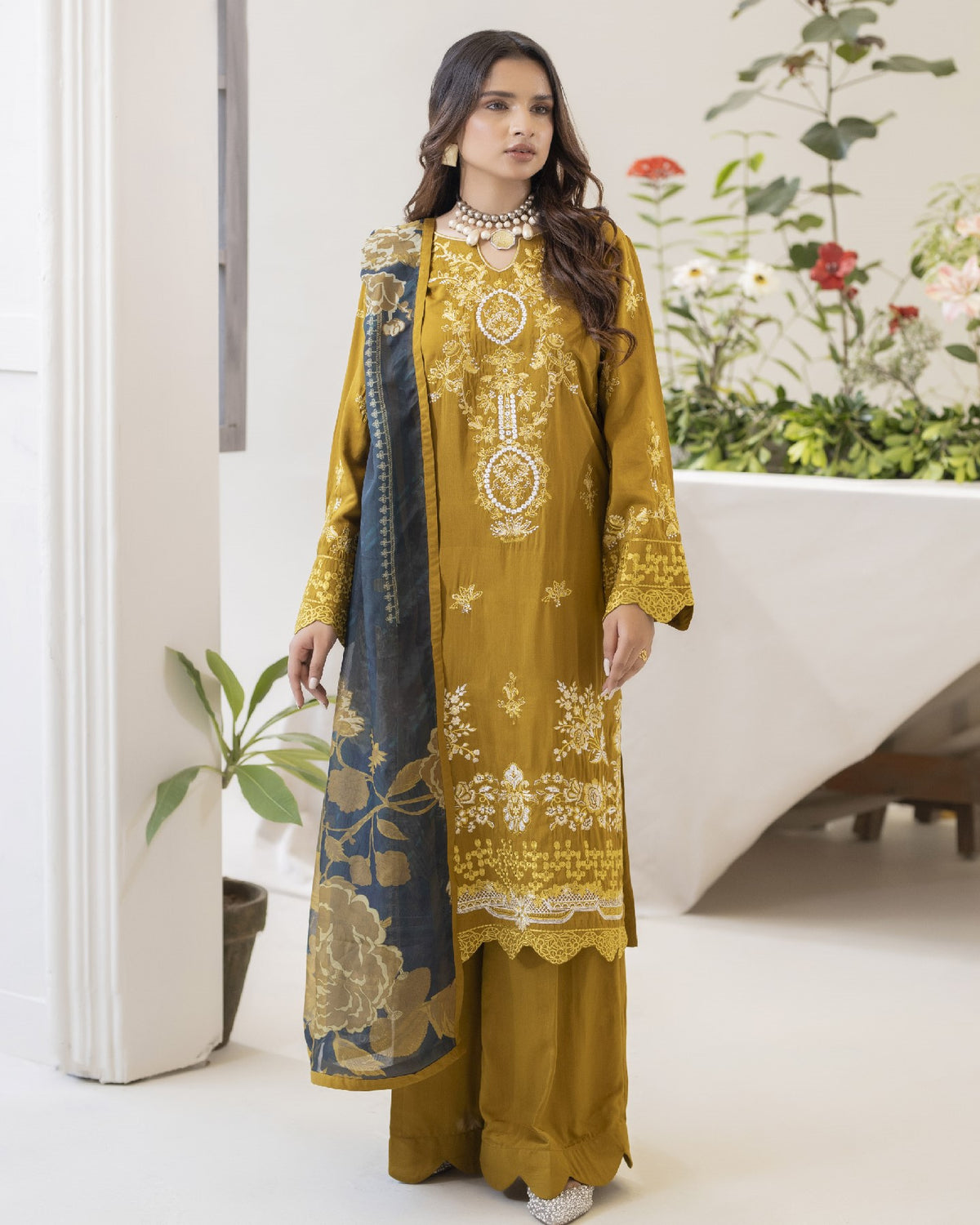 Woman wearing a mustard yellow embroidered outfit with a matching dupatta in a minimal indoor setting.