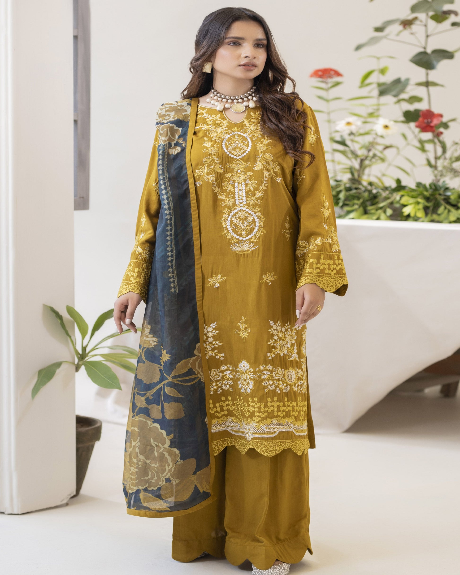 A woman wearing a yellow embroidered shirt with floral patterns, a blue dupatta, and matching trousers.