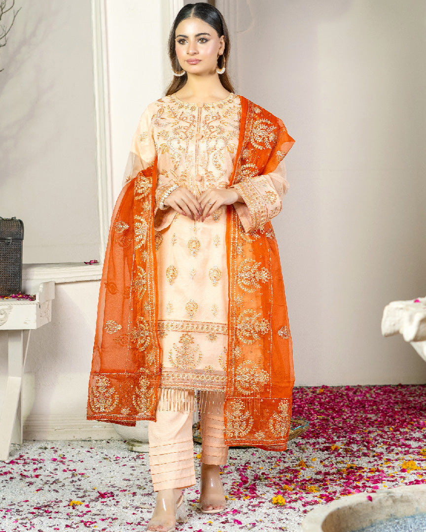 A woman wearing a traditional outfit consisting of a beige shirt with embroidery, an orange dupatta, and a matching beige trouser. The outfit features intricate embroidery details.