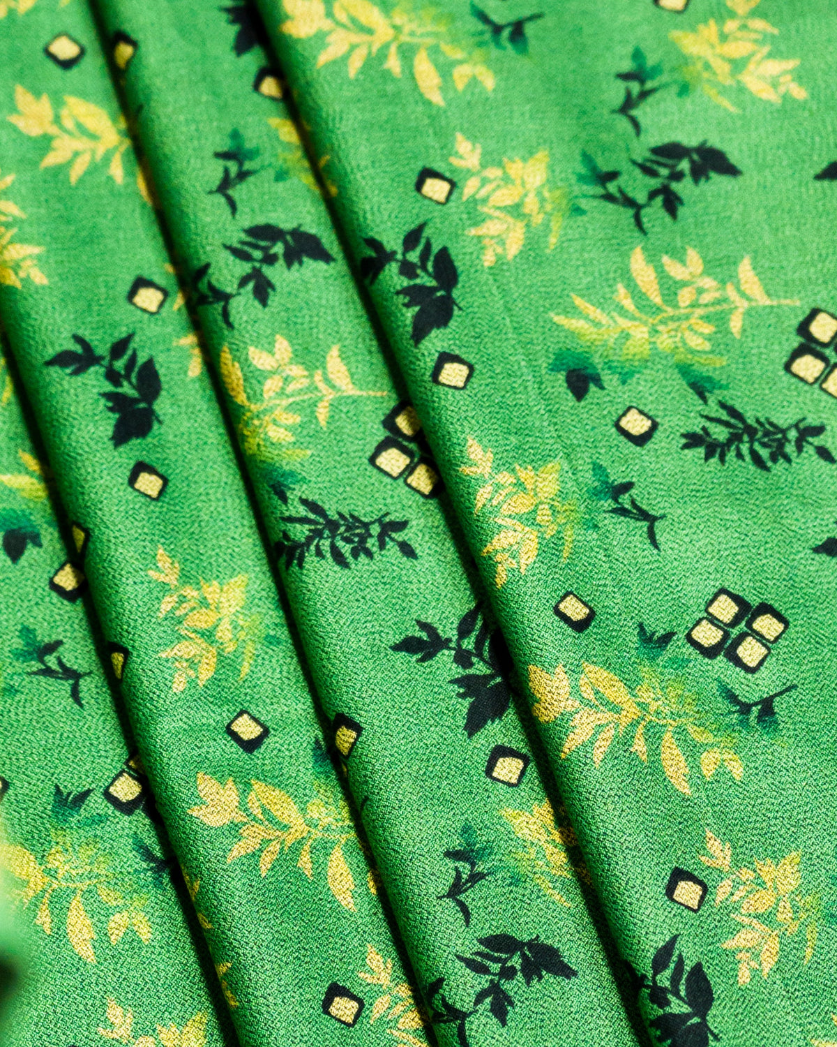 Green patterned fabric folded in a stack, featuring a black and yellow botanical print with small diamond motifs, shown on a white surface next to dark wooden objects.