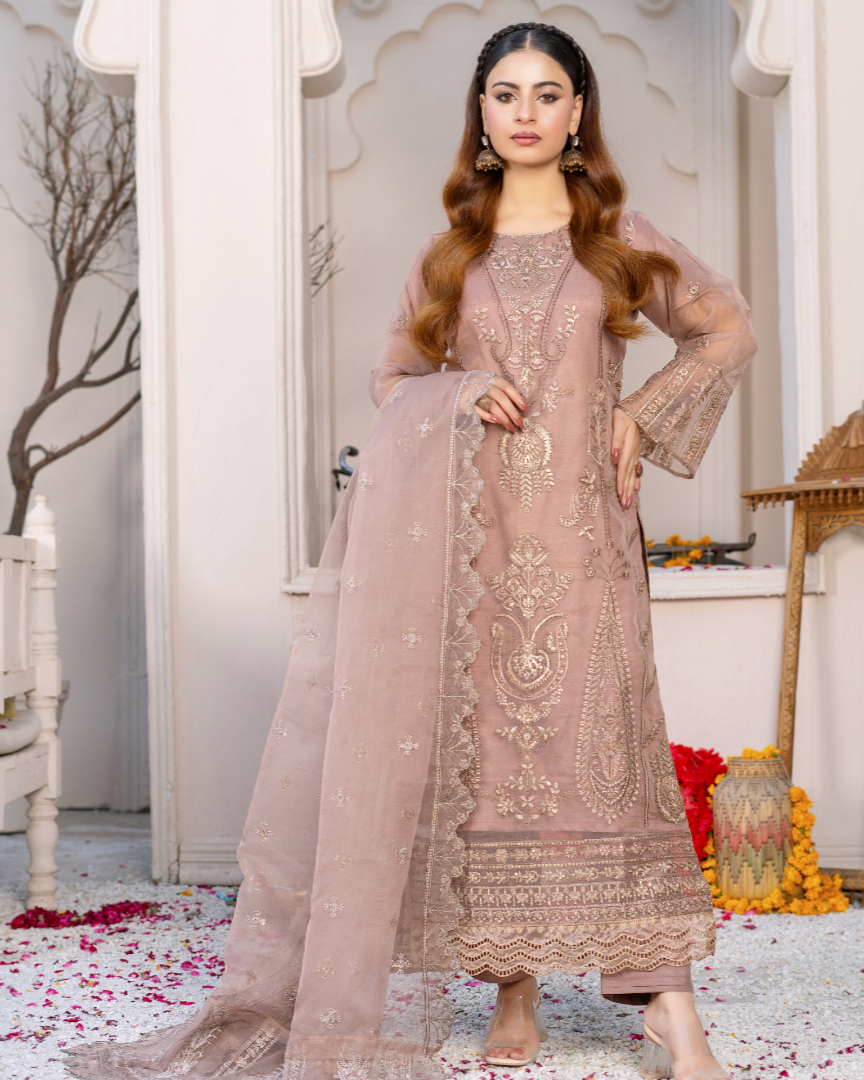 A woman wearing a traditional embroidered dress in a beige color with floral patterns, paired with trousers, standing in a decorative setting.