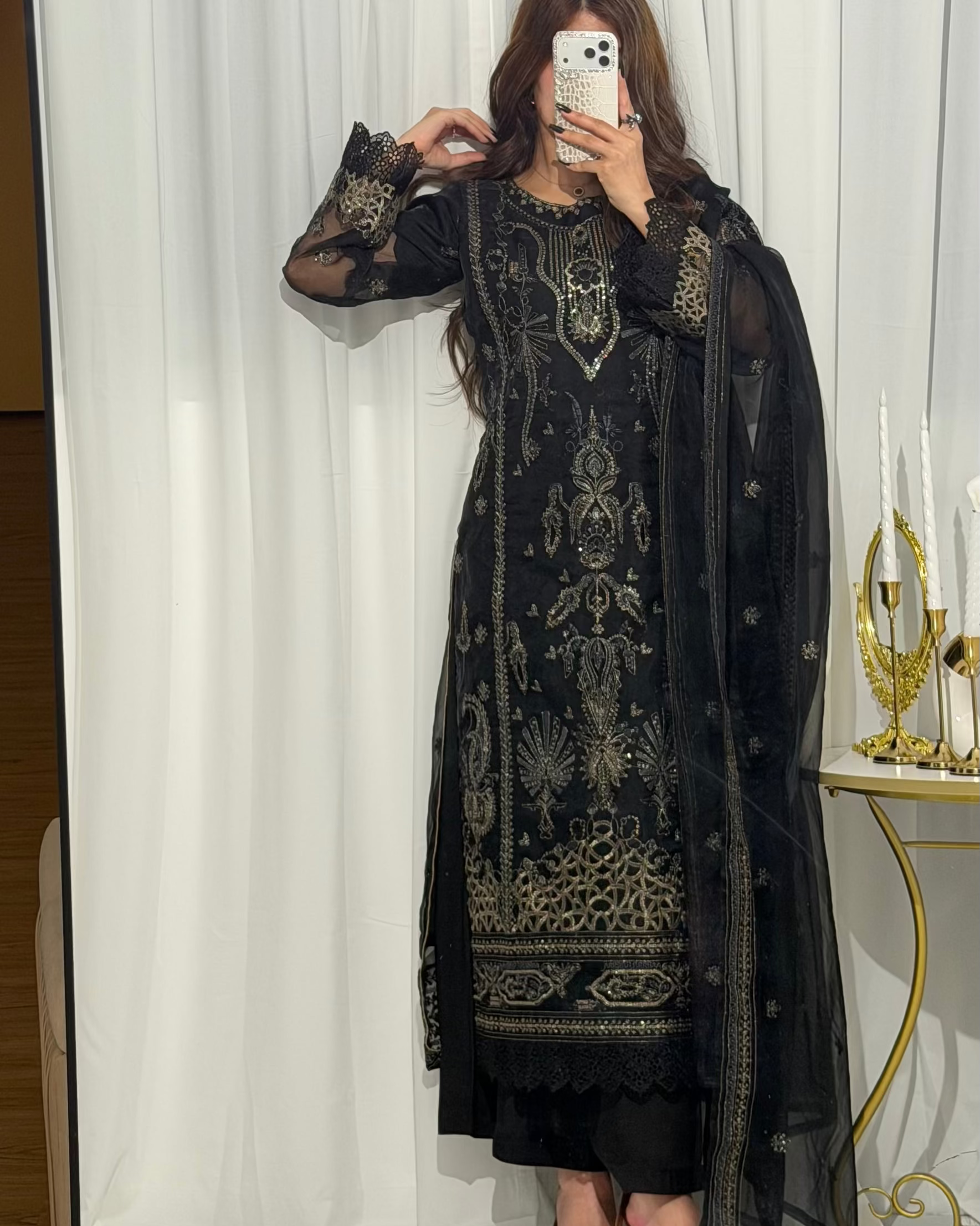 Person wearing a black embroidered traditional outfit in front of a white curtain.