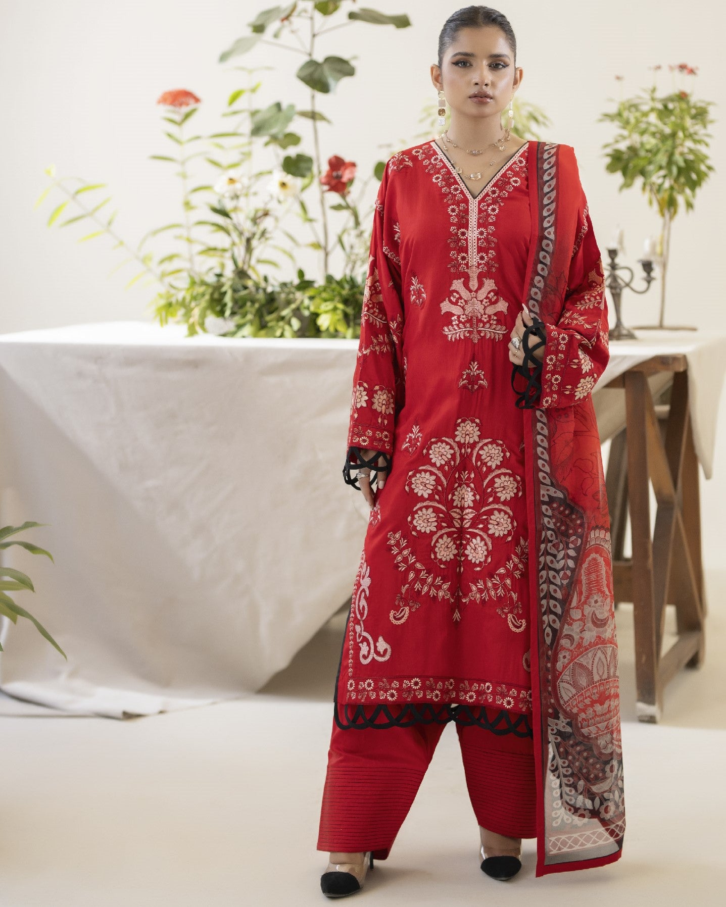 Woman wearing a red embroidered traditional outfit with a white background and plants.