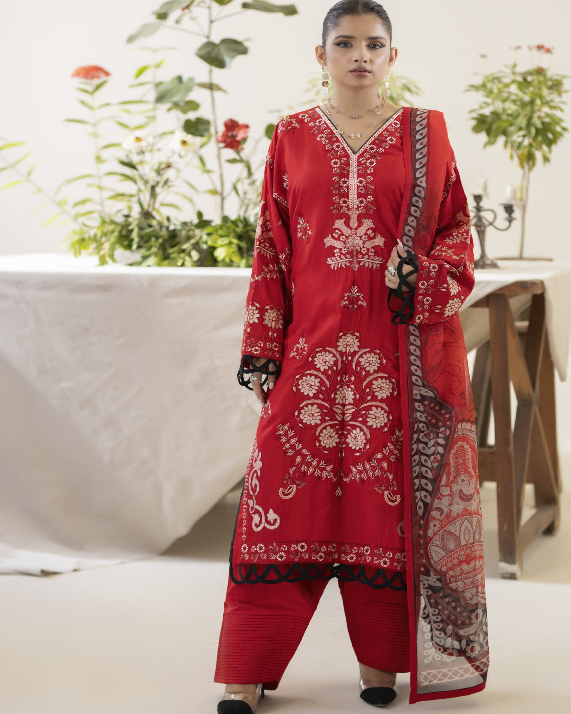 A woman wearing a traditional red embroidered shirt with a dupatta and trousers, standing in a setting with plants and a table in the background.