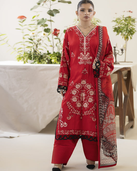 A woman wearing a traditional red embroidered shirt with a dupatta and trousers, standing in a setting with plants and a table in the background.