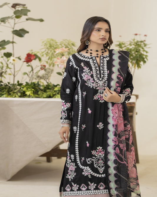 A woman wearing a traditional black embroidered shirt with floral patterns, paired with a digital printed silk dupatta in pink and green shades and viscose dyed trousers, standing in a floral setting.